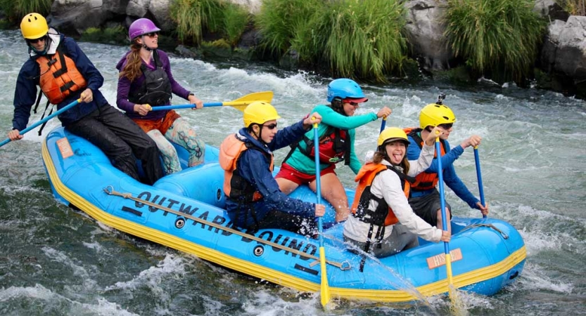 a group of students wearing helmets and life jackets paddle a blue raft through whitewater